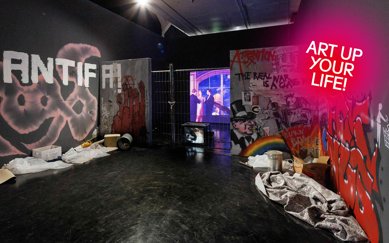 Installation in einem Ausstellungsraum mit politischen Graffiti, Absperrgitter, Projektionsbildschirm und verstreuten Materialien auf dem Boden.