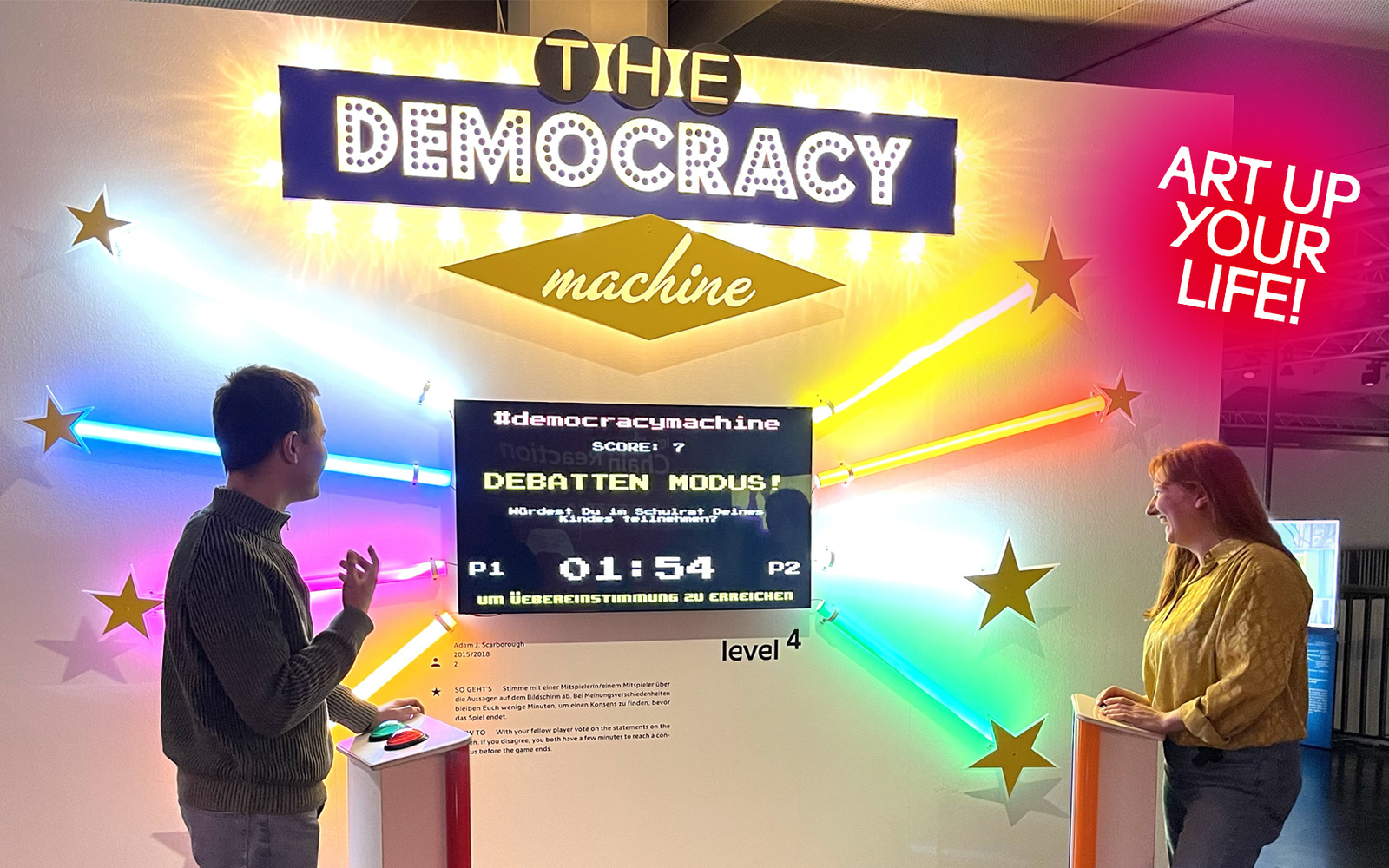 Two people stand facing each other in front of the “Democracy Machine.”