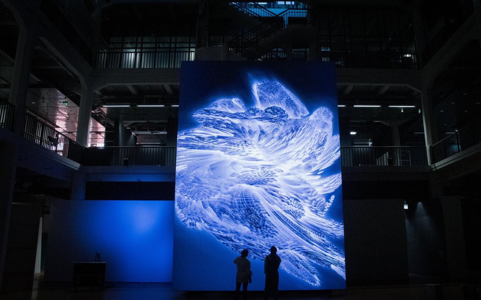 Zwei Silhouetten stehen vor einem riesigen Bildschirm, auf dem ein abstraktes Kunstwerk zu sehen ist.