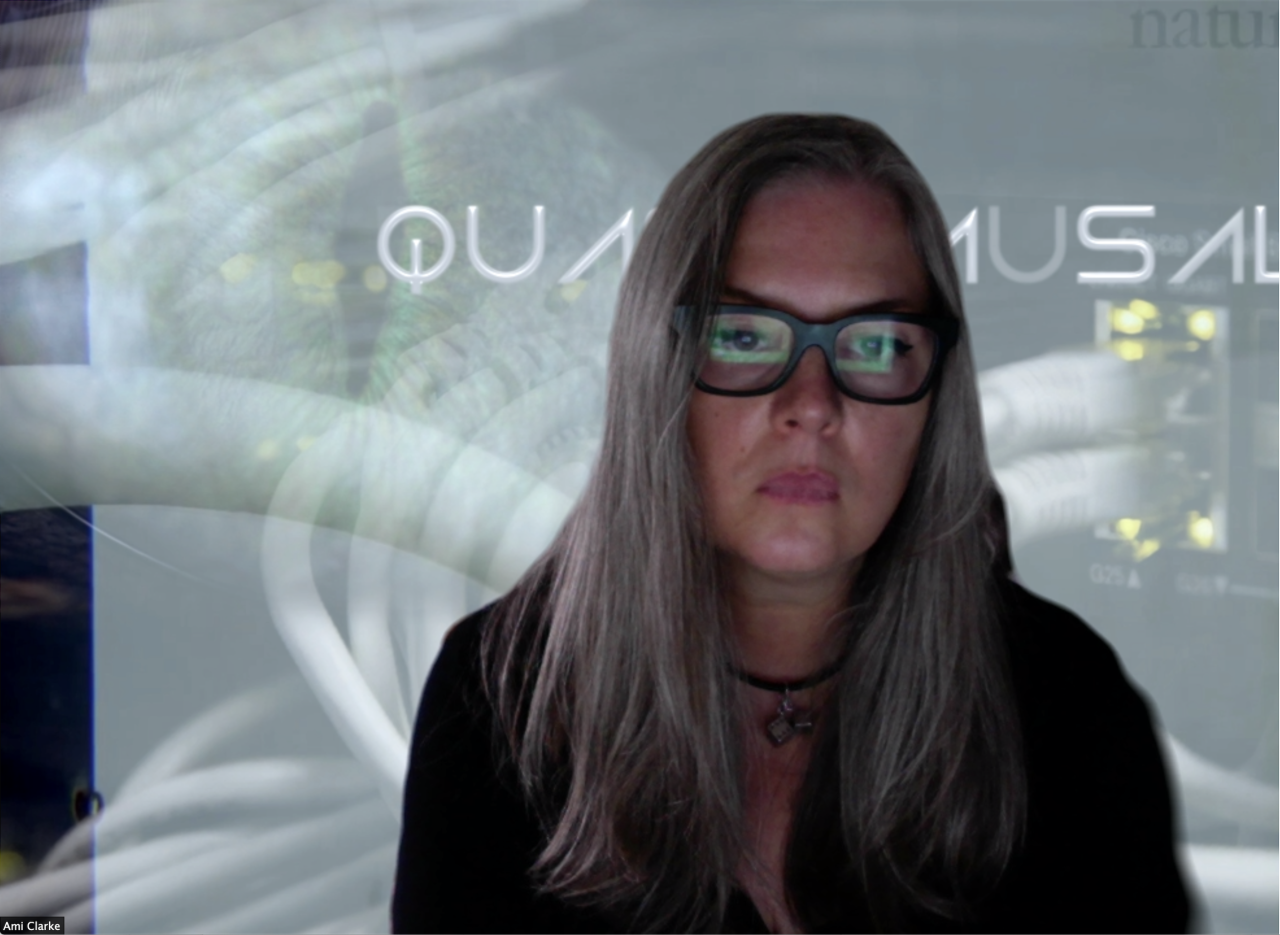 Ami Clarke, a woman with long hair and glasses with a thick, square frame, looks at a screen. This screen is reflected in her glasses.
