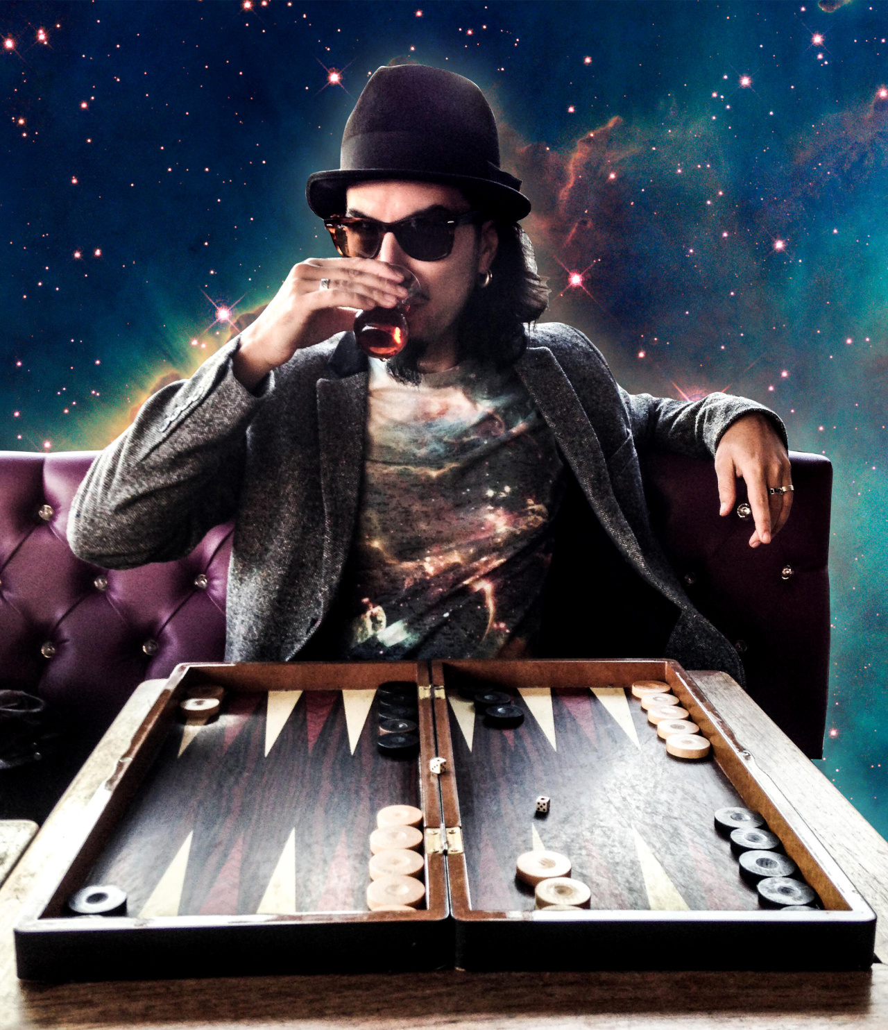 Memo Aktens is sitting on a violet velvet couch and in front of a backgammon game, drinking turkish tea. The background is a colage of the universe. 