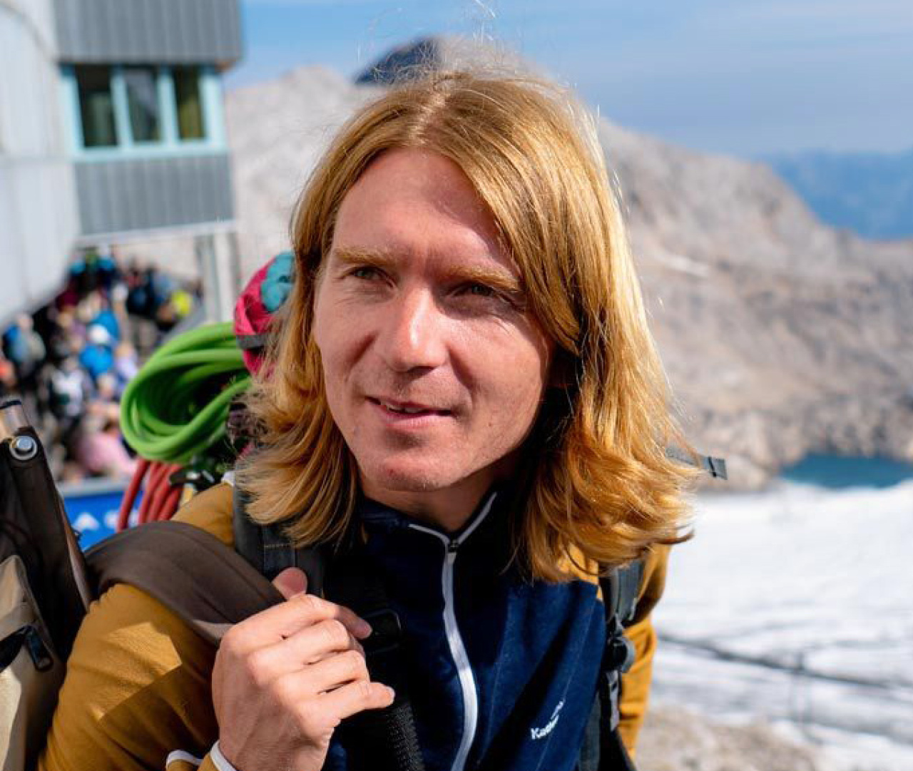 The picture shows the artist Markus jeschanuig. He is pictured outside and seems to be on a hike on a mountain top. He wears open shoulder-length blond hair and looks left out of the picture.
