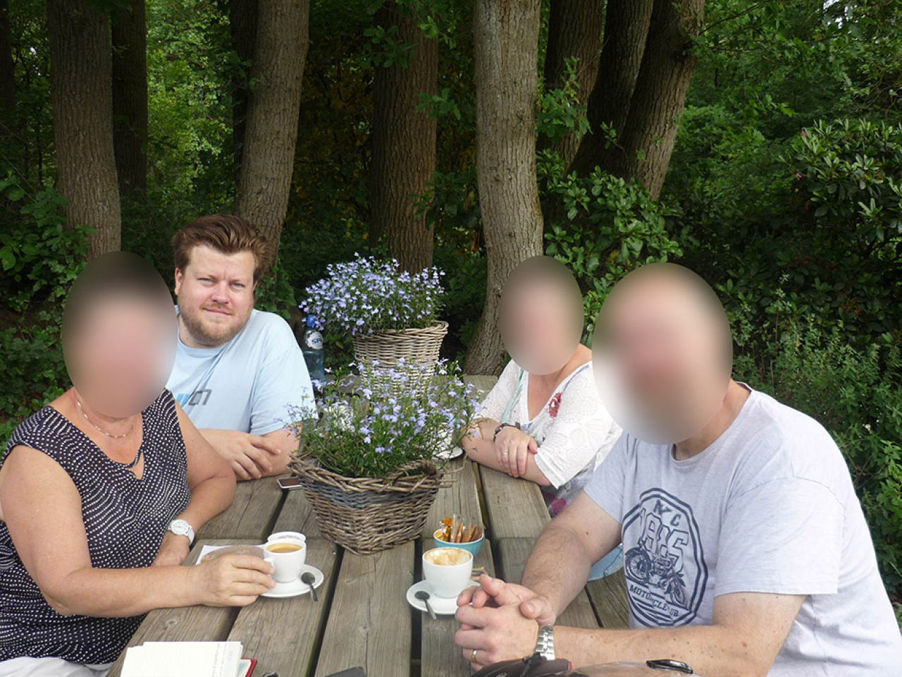 Auf dem Foto sieht man den Künstler Constant Dullaart, der mit drei zensierten Personen an einem Holztisch im Freien sitzt.