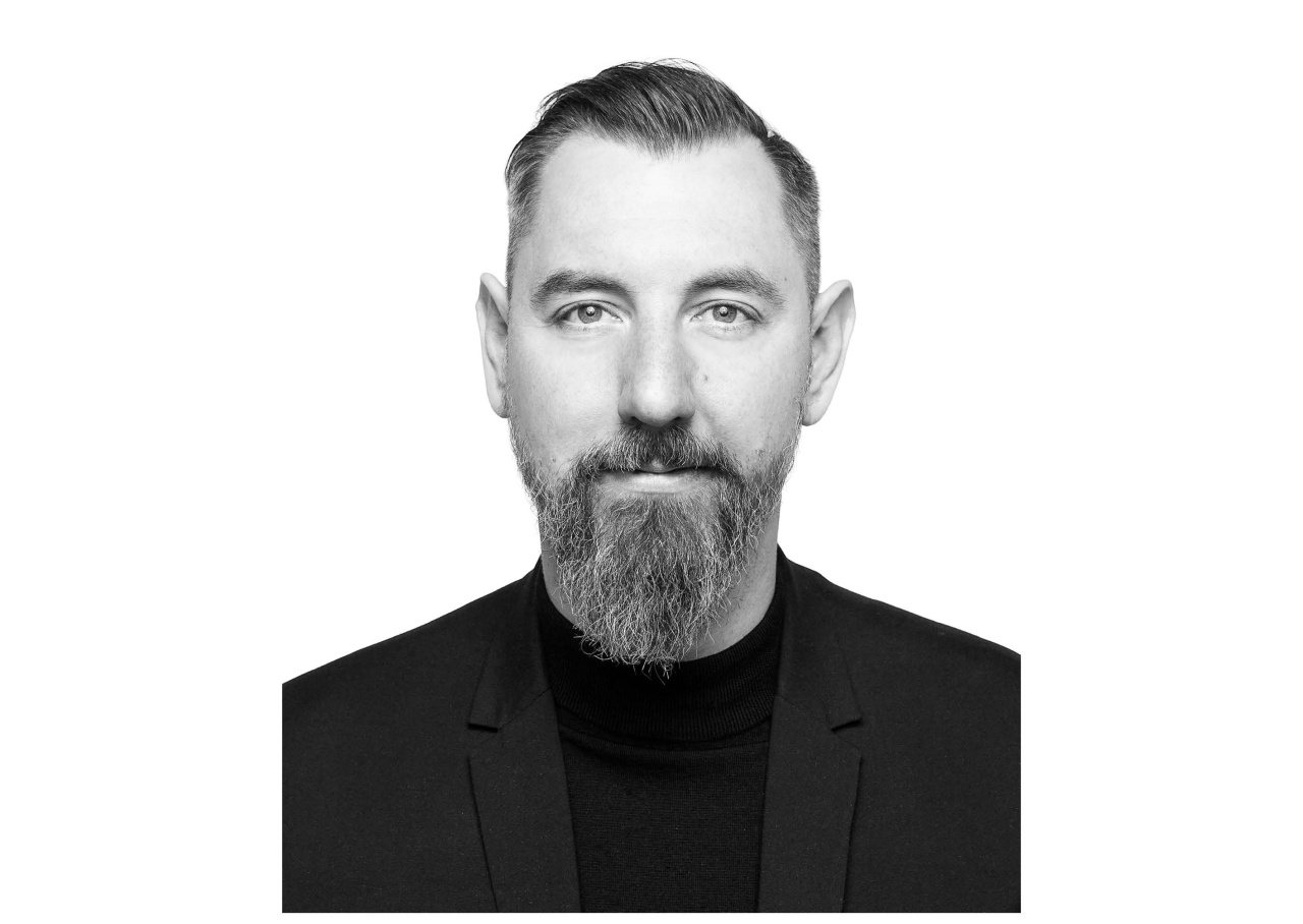 Zu sehen ist ein Porträt von Stephan Schwingeler. es ist ein schwarz weiß Foto und zeigt ihn ab der brust aufwärts. Er trägt einen Bart und ein schwarzes T-shirt und schaut frontal in die Kamera.