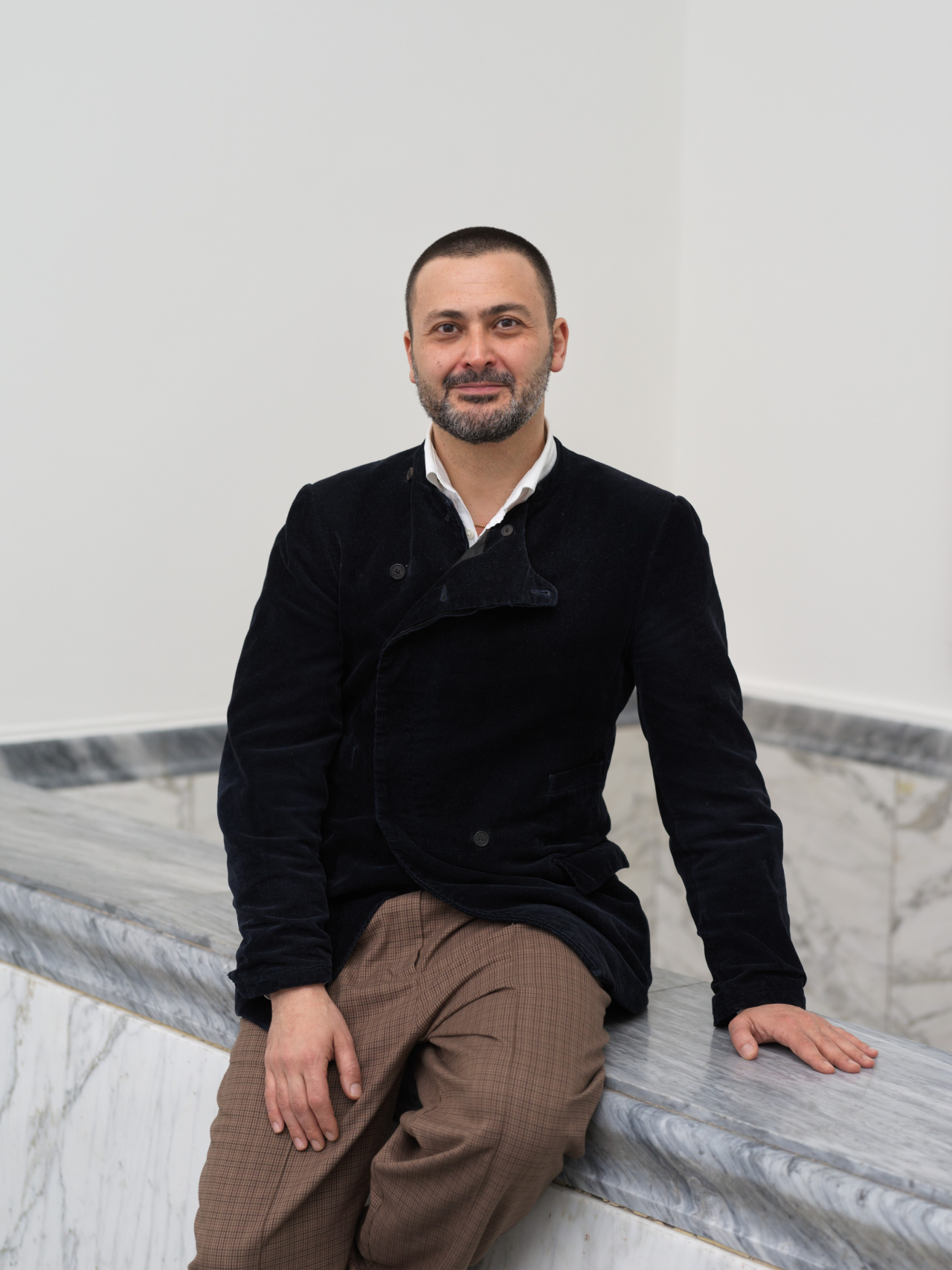 Portrait of Misal Adnan Yildiz sitting on a marble wall in front of a white wall