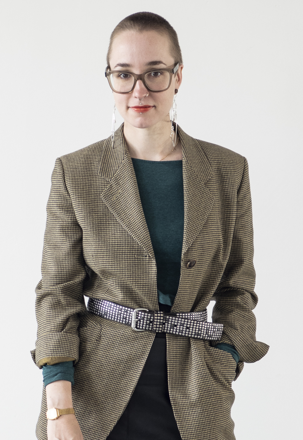 A young woman with glasses stands in front of a white background and wears a beige plaid coat.