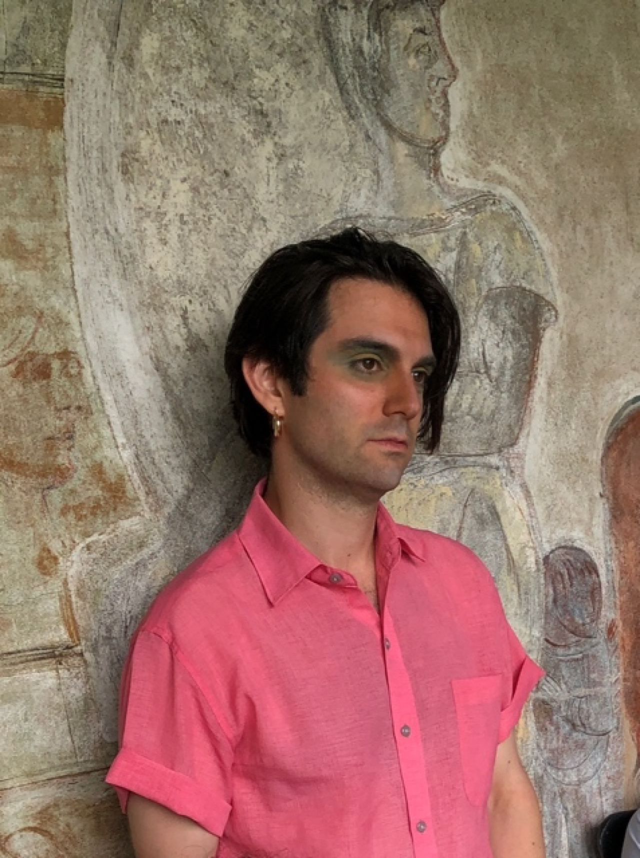 A black-haired man in a pink shirt can be seen leaning against a stone wall and looking out of the picture to the right.