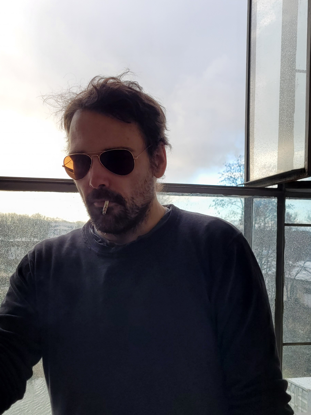 Christian Schlaeffer smoking in front of a window pane wearing a black sweater and sunglasses
