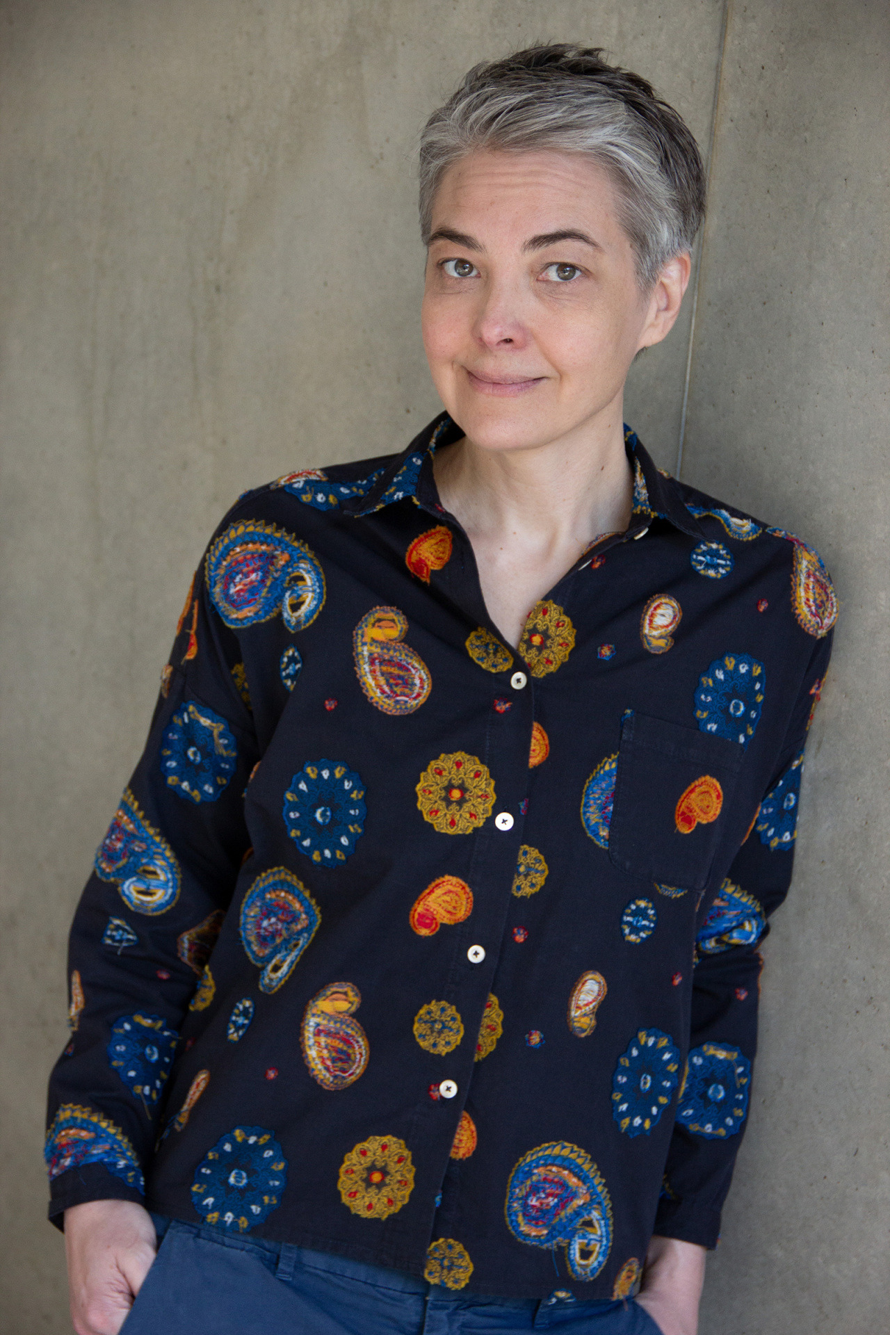 A woman with short hair and a colorful blouse leans against a concrete wall