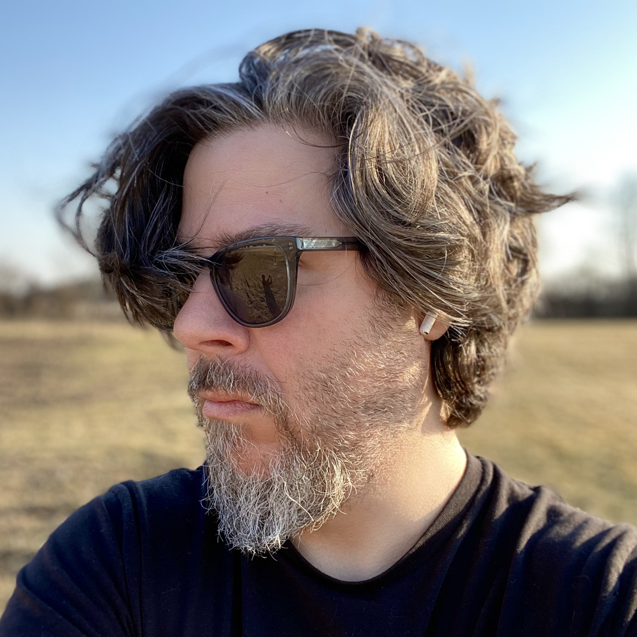 Das Bild zeigt einen Mann mit dunklen Haaren und grauem Bar von der Seite. Eine Sonnenbrille verdeckt seine Augen. Im Hintergrund ist ein Feld und blauer Himmel.