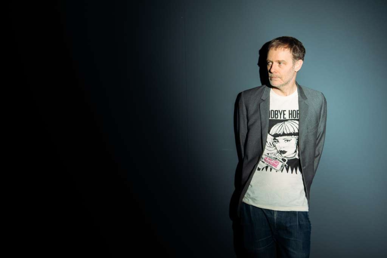 A person is standing with their arms crossed behind their back against a grey background.