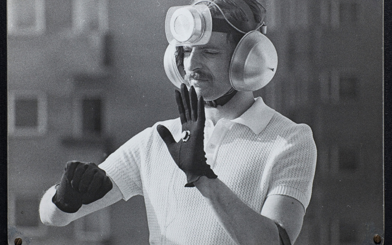 Das Schwarzweiß-Photo zeigt einen Mann mit einer technischen Installation auf dem Kopf, seine Ohren sind mit Schallschützern bedeckt.