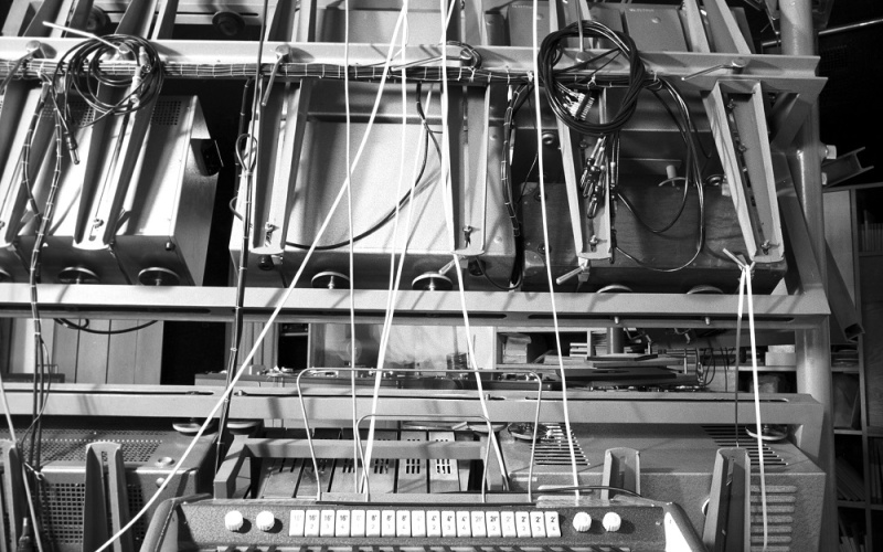 A black and white picture shows an electric piano in front of cables and computers. 