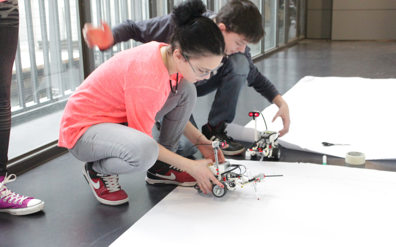 Two kids are sitting on the floor in front of a big sheet of paper and are setting two lego-robots on the paper.