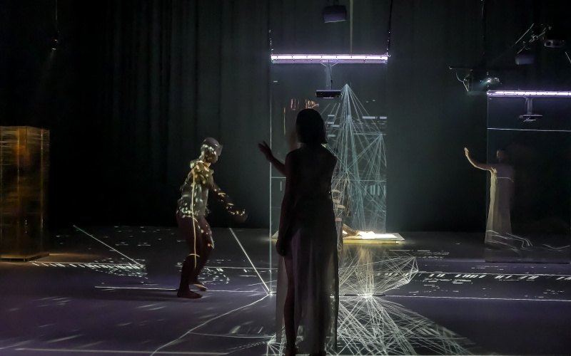Three dancers stand on the stage, it is dark. In the middle of the stage is an installation of laser light. On the body of the left dancer is a projected play of light.