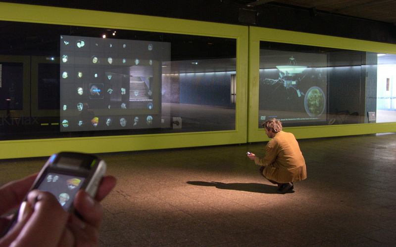 Auf die Glaswände des Ausstellungsraums ZKMax wird »SpacePlace« projiziert. Davor sieht man Personen mit Mobiltelephonen, die mit der Installation kommunizieren.