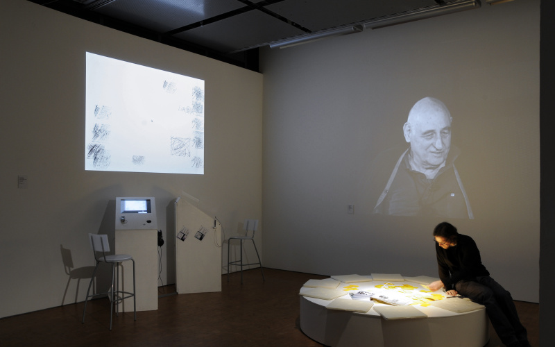  On the back wall a black and white projection showing Kurd Alsleben. In the foreground a man on a round pedestal, reading.