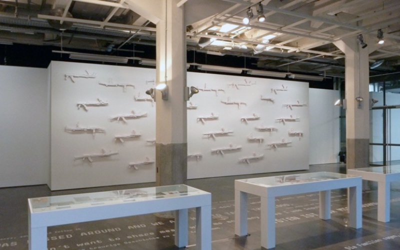 Blick in die Ausstellung »Symbiosis«: Im Vordergrund: weiße Vitrienen, im Hintergrund an der Wand: weiße Gewehre 