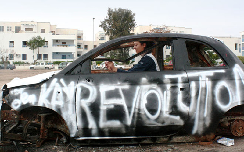 A woman sitting in a non-functioning car. On the driver's side was sprayed with white color "revolution".