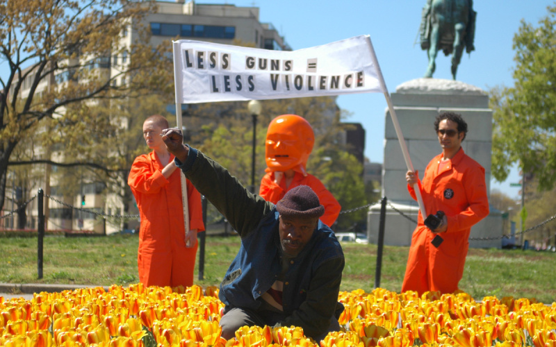 Ein Mann im Blumenbeet hebt seine rechte Faust als Zeichen des Protests. Hinter ihm drei Männer mit einem Transparent.