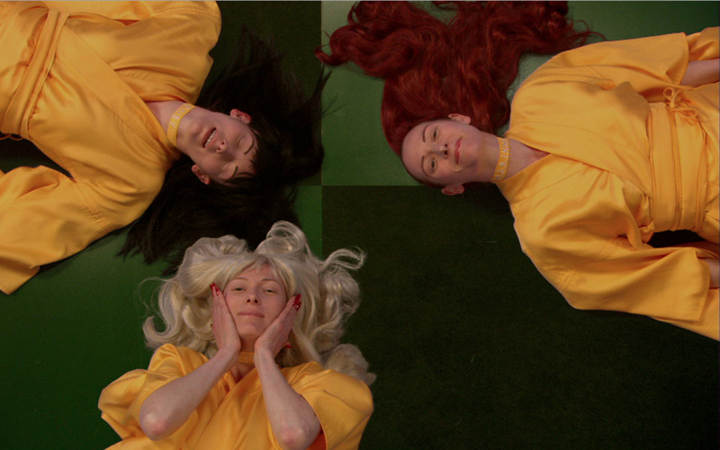 three women, each with blond, black and red hair color lie with their heads together on the floor and looking into the camera