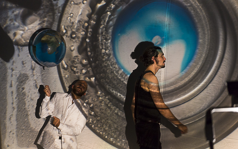 A man dressed in black standing in profile in front of a large silver circle element whose center is bluish. Behind him a man dressed in white holding a balloon in his hands.