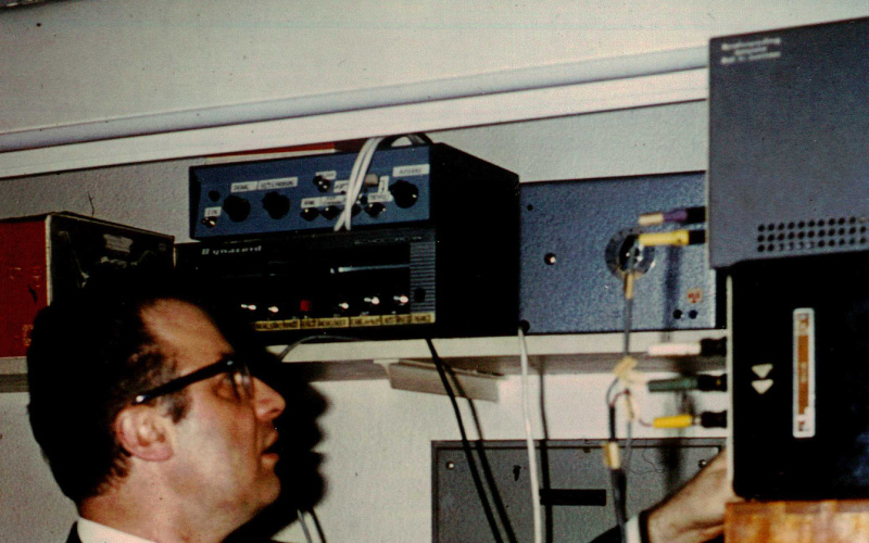 Ein Mann im schwarzen Anzug und schwarzer Hornbrille bedient diverse elektronische Gerätschaften.