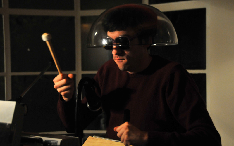 Ein Mann mit Sonnenbrille und durchsichtigem Lampenschirm auf dem Kopf, der einen Drumstick in seiner rechten Hand hält.