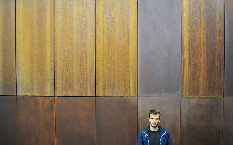 Ein junger Mann mit blauer Sportjacke steht vor einer Holzwand