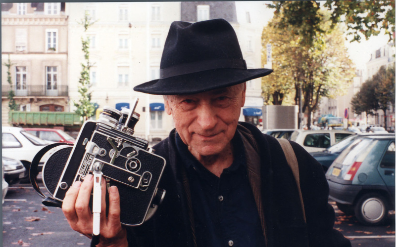 Jonas Mekas with a hat on his head and a camera in hand