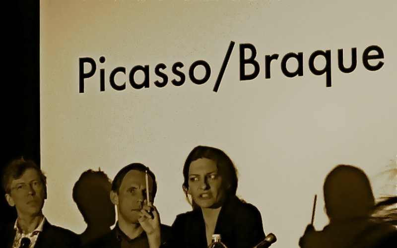 Von links nach rechts: Zwei Männer und eine Frau an einem Tisch. Sie redet und hält dabei einen Bleistift in ihrer rechten Hand. Auf dem Tisch stehen Namensschilder. Im Hintergrund ist 'Picasso/Braque' zu lesen.