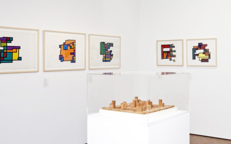 Ein Blick in die Ausstellung »Hiroshi Kawano«: Ein Raum mit sieben Gemälden mit geometrischen Mustern, in der Mitte eine abstrakte Holzskulptur.