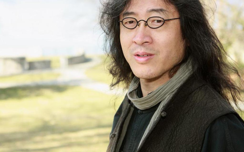 A man with glasses and long hair stands in front of a tree