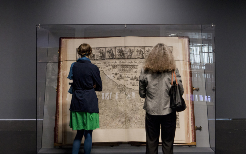 The Klencke Atlas (1660) is one of the largest atlas in the world. Aerated he stands in a glass structure that protects it. Two women seen from behind looking at it.