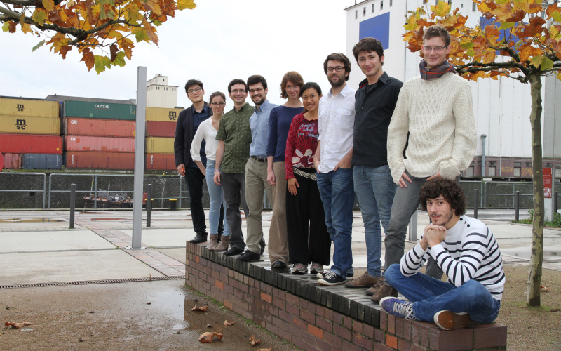 Nine people are standing on a small wall. A tenth is sitting. In the background a harbor area.