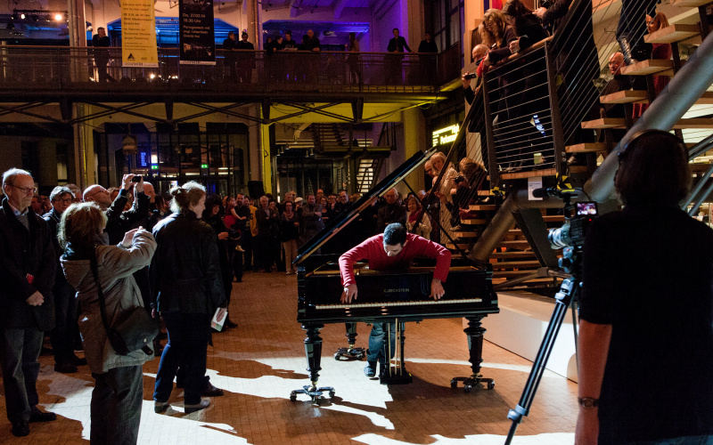 A man stands in a piano. From this position he plays the piano. Around him are many people who are watching him.