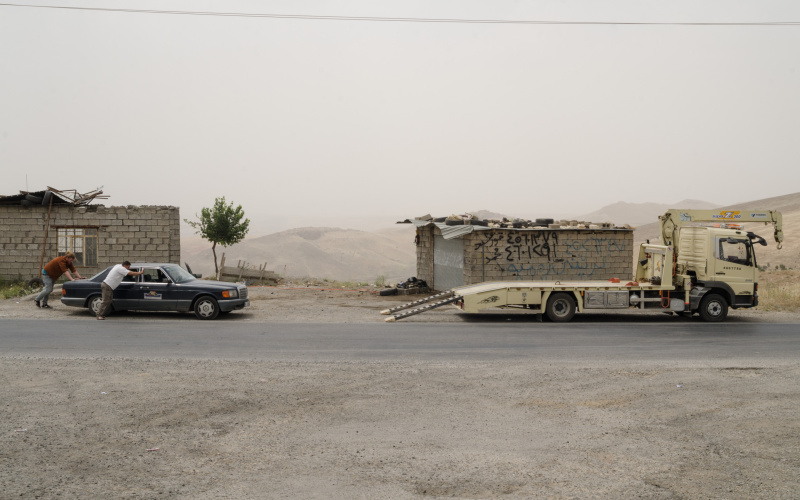 Zwei Männer schieben einen Mercedes in Richtung eines Abschleppautos. Hinter ihnen ruinöse Gebäude und eine karge hügelige Landschaft.