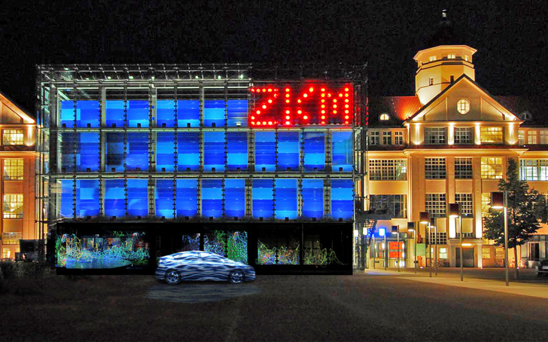 Ein Auto steht vor dem blauen Kubus. Rechts oben in großen, roten leuchtenden Lettern: ZKM