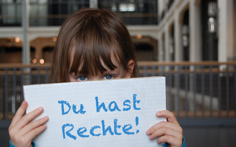 A little girl holding a placard in front of her face: "Du hast Rechte!"