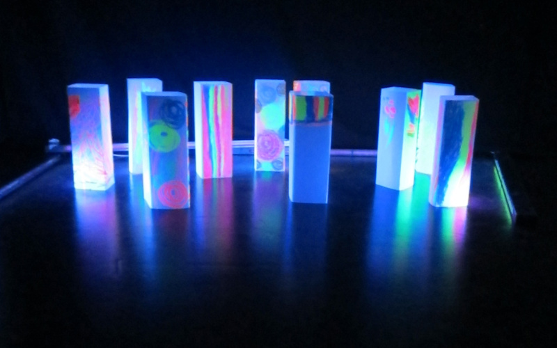 Numerous, with bright colors painted blocks are glowing in the neonlight. They stand in front of a black background and resemble a skyline.