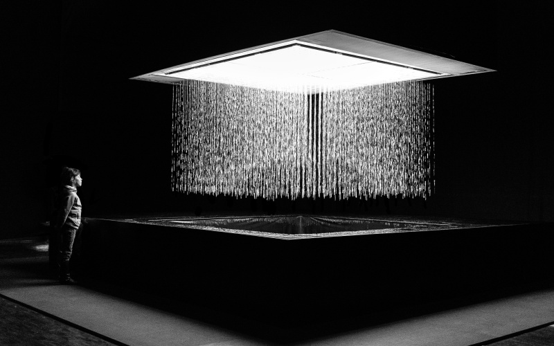 Schwarzweißaufnahme der Installation 3D WATERMATRIX, die Wasser formgebend von der Decke zum Boden fallen lässt. Eine Person betrachtet die Installation.