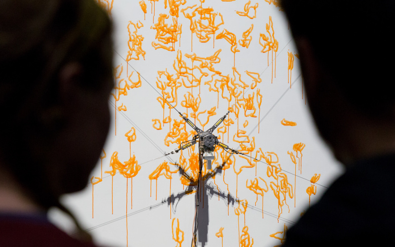  Two silhouettes looking at the computer-controlled robot painting with a yellow paint on a canvas.