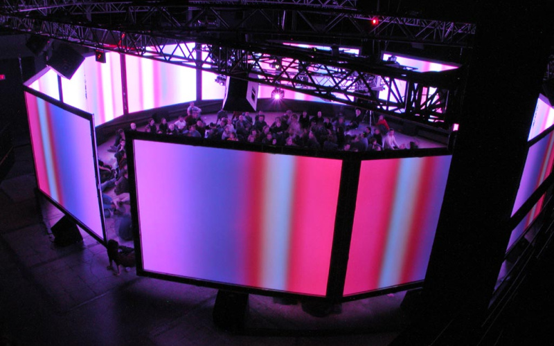 Circularly arranged, colorful illuminated screens in the middle of which a group of people is sitting.