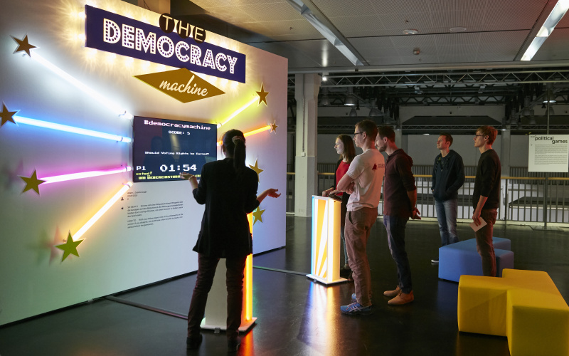 A screen, colorful light sticks and the neon sign The Democracy Machine are attached to a white wall. Two visitors stand in front of the controls of the performative sculpture by Adam Scarborough. More visitors are watching. 