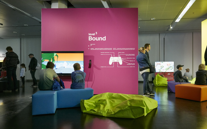 Two visitors sit on a blue bench in front of a magenta-coloured wall with a screen attached to it. Also in the foreground is a neon green beanbag. Right and left in the background, more visitors. 