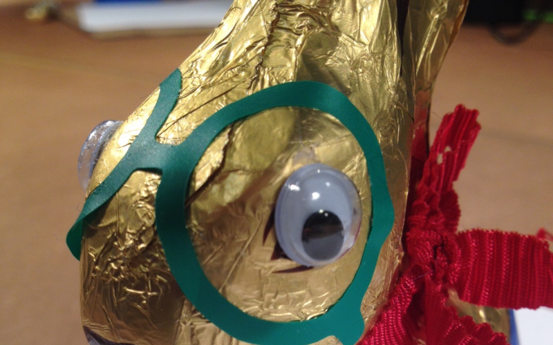 A golden chocolate Easter bunny looks into the camera with its wobbly eyes glued on. Beside the eyes a green sticker similar to glasses is glued on the packaging of the Easter bunny.