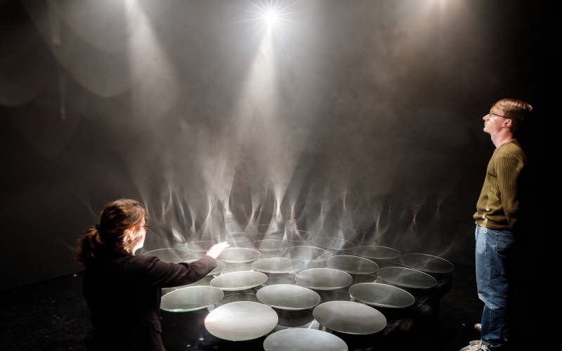 Two visitors look at the columns of smoke, which are made visible by the focusing mirrors in interaction with the light.