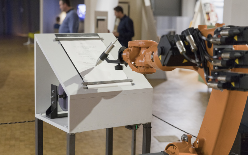 Ein oranger Roboterarm schreibt mit einem großen silbernen Stift auf Papier