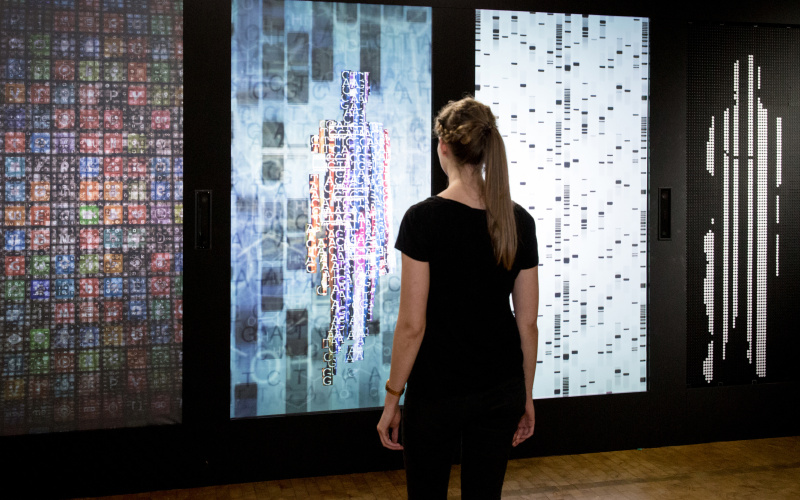A woman stands in front of the installation YOU:R:CODE, a multi-channel projection which she presents in data form.