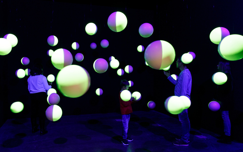 In einem dunklen Raum hängen halbseitig-fluoreszierende Styroporkugeln von der Decke mit denen drei Besucher interagieren